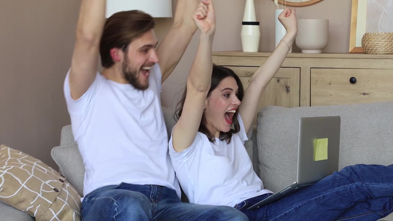 Pareja joven emocionada celebrando el éxito mientras usa el portátil en el sofá