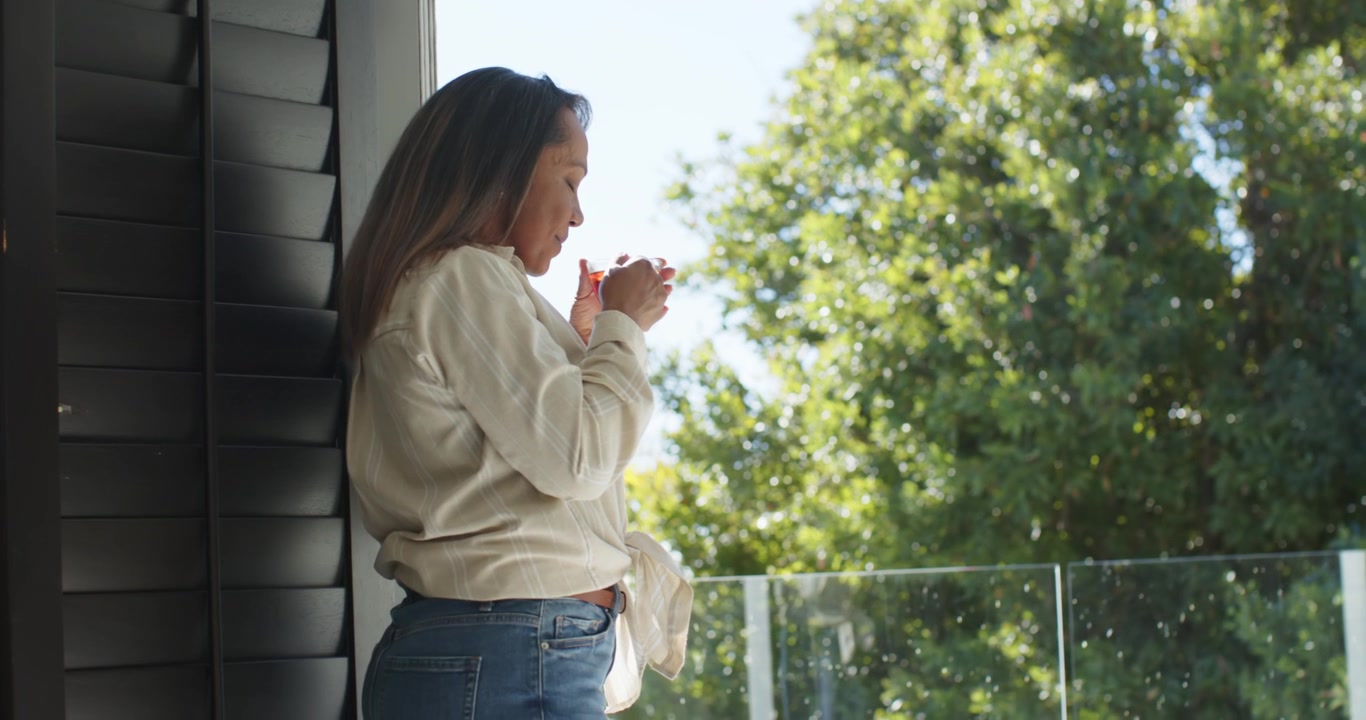 Donna asiatica matura in piedi sul balcone che tiene una tazza, guardando in basso e inalando l'aroma dalla tazza.