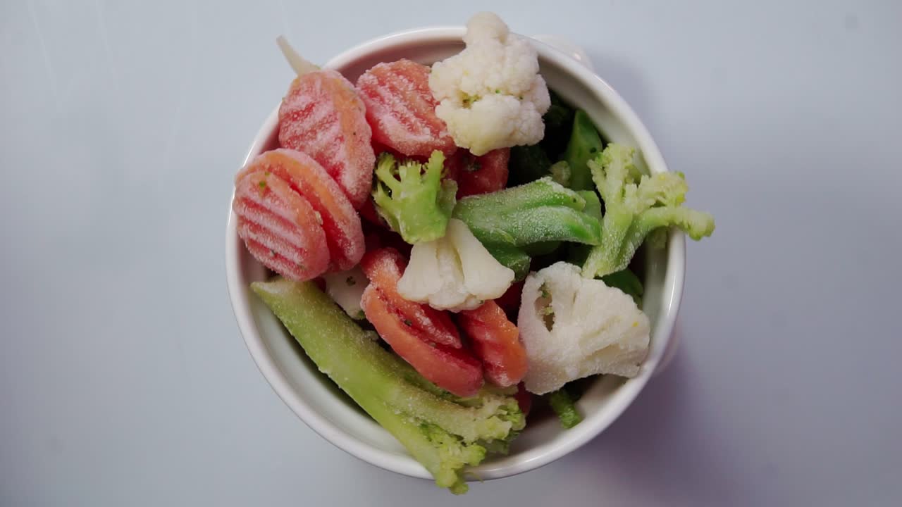 A bowl with frozen mixed vegetables rotating on a white surface under soft daylight, cinematic look and detail