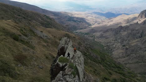 Mountain-aerial-descends-to-two-people-standing-on-narrow-rock-fin