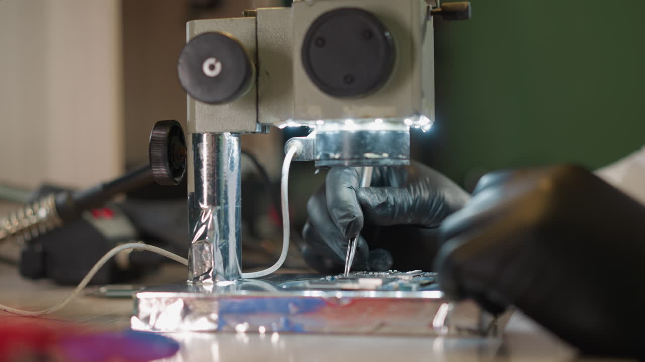 검은 장갑을 입은 기술자의 손이 도구를 사용하여 현미경 아래 회로판에서 정확하게 작업하는 클로즈업 뷰; 테이블에는 다양한 전자 장비가 장착되어 있습니다.