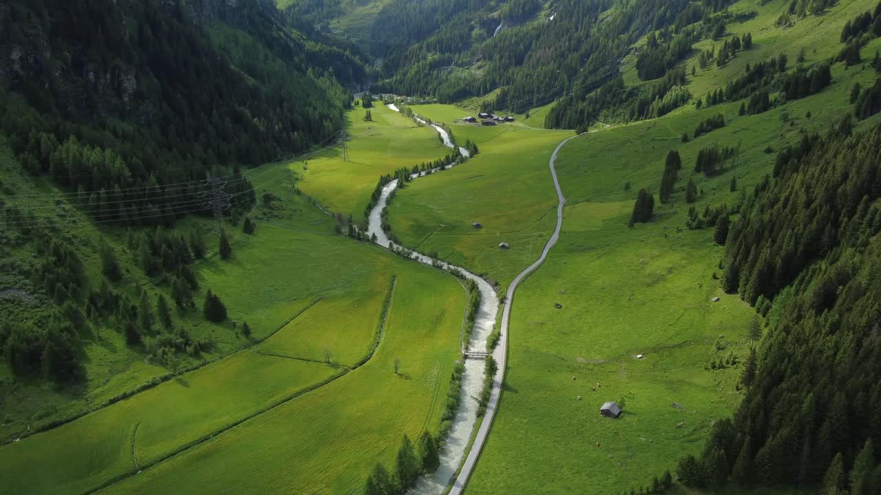 Aerial shot of a winding river through vibrant green alpine meadows and forests.