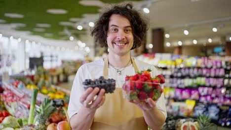 Portrait-of-a-happy-brunette-supermarket-worker-holding-small-boxes-of-blueberries-and-strawberries-in-his-hands