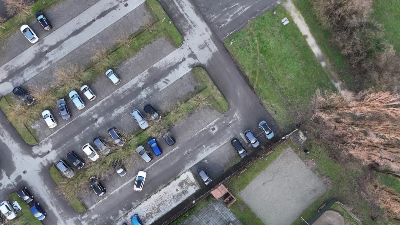 From above, a white car sits in a grid-style parking lot bordered by grassy dividers and leafless trees, with tiled rooftops and pathways framing the urban layout