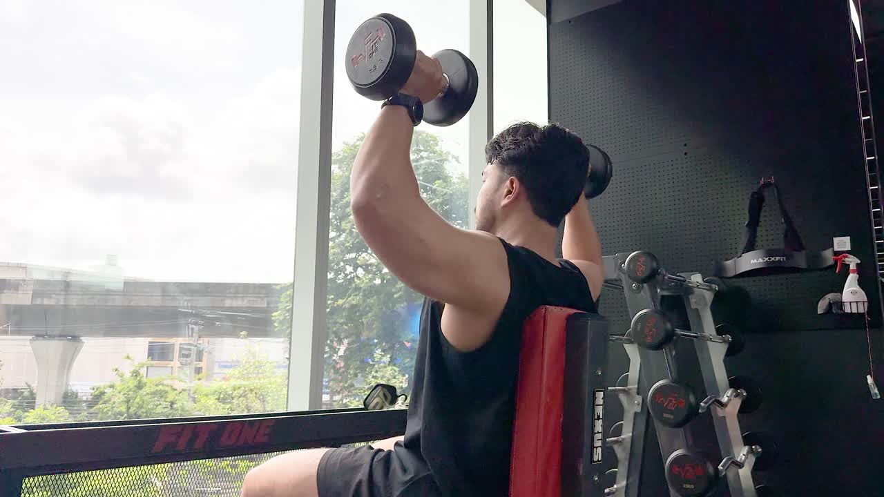 A person performs a dumbbell shoulder press in a well-lit gym with large windows, showcasing strength and fitness