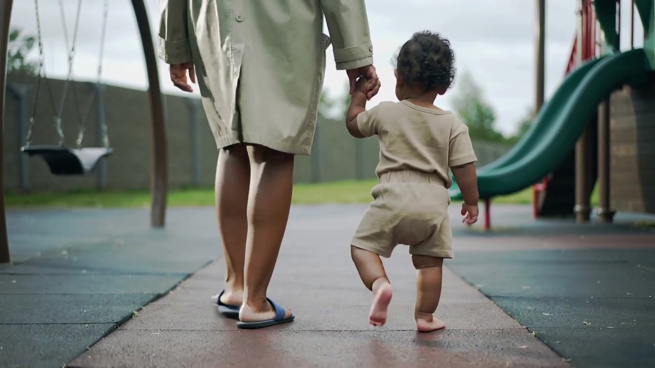 Guiding adult in coat, sandals holding toddler's hand, practising steps at park to slide and swings