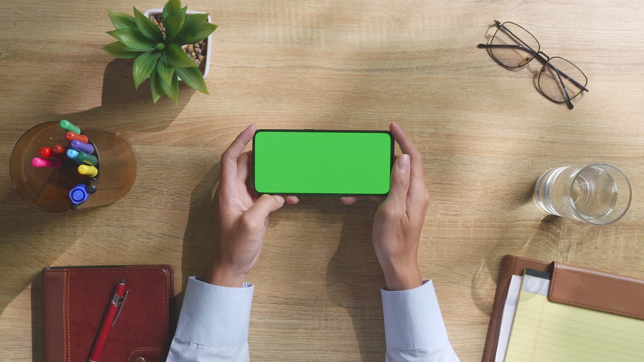 vista de arriba hacia abajo de una computadora portátil con pantalla verde simulada pantalla chromakey en un escritorio de oficina de madera al lado de la libreta con bolígrafos, gafas y un vaso de agua.