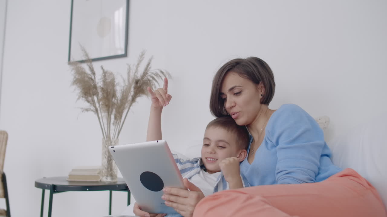 mamá e hijo mirando la pantalla de la tableta tendida en una cama blanca. juega juegos con tu hijo en tu tableta y mira videos divertidos.