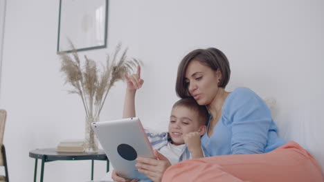 mamá e hijo mirando la pantalla de la tableta tendida en una cama blanca. juega juegos con tu hijo en tu tableta y mira videos divertidos.