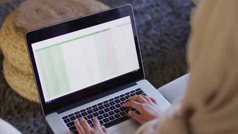 over shoulder video of biracial woman in hijab at home working on laptop, spreadsheet on screen