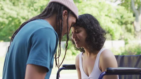 Happy-biracial-couple-touching-heads-in-garden,-woman-sitting-in-wheelchair,-man-with-dreadlocks