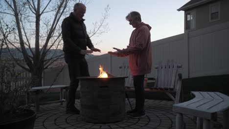 senior man joins his wife as the warm their hands at a backyard fire pit