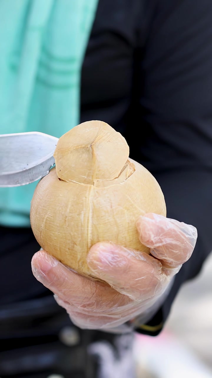 Venditore guantato affetta abilmente la parte superiore di una noce di cocco con un machete, preparando una bevanda tropicale in un mercato all'aperto.