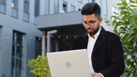 Happy-Indian-businessman-typing-working-remote-on-wireless-laptop-computer-browsing-online-outdoors