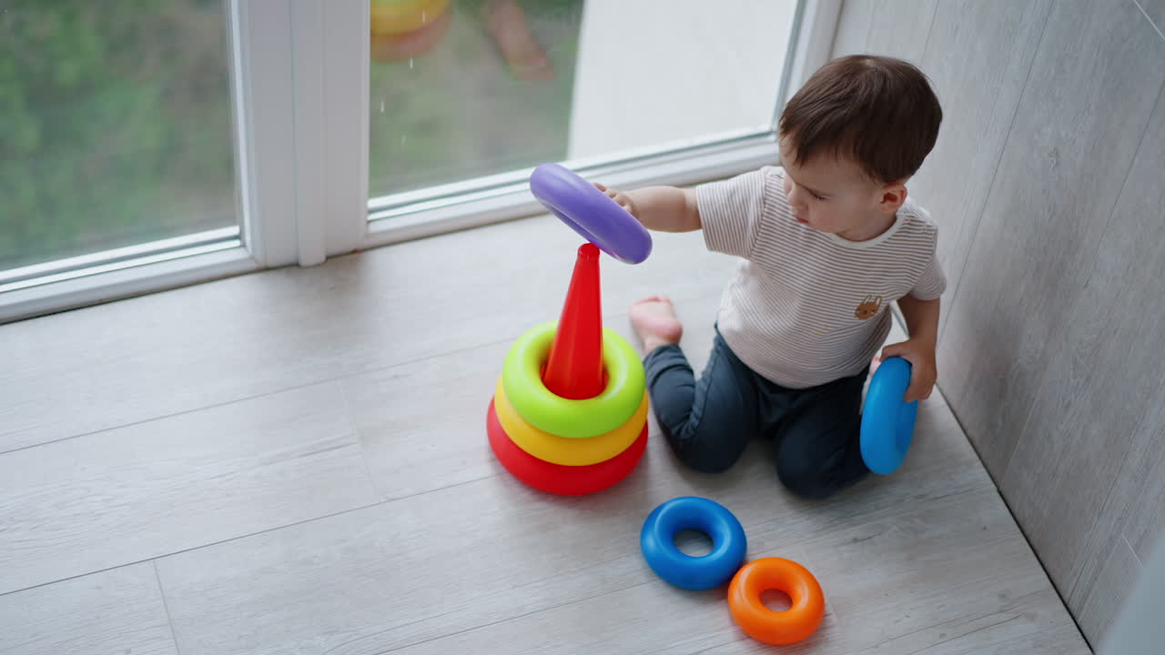 Adorable Caucasian toddler sits on his knees on the floor. Child constructs the pyramid on the balcony. View from top.