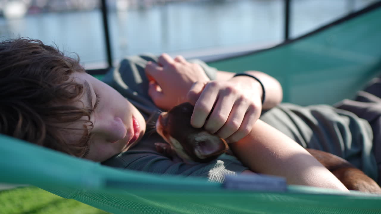 Teenage boy lying peacefully in green hammock, gently stroking small brown chihuahua near marina waterfront with sailboats and soft afternoon sunlight