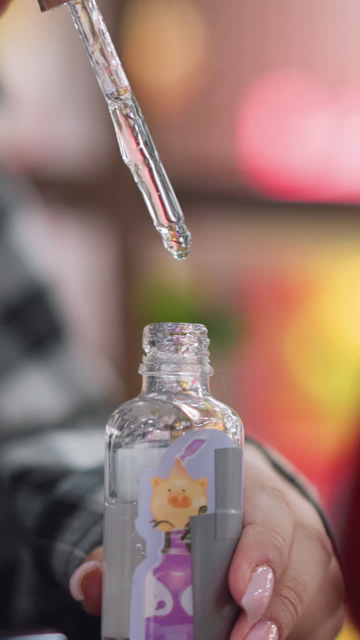 Closeup glitter serum dropper above bottle, womans hand with pink manicure holding decorated glass bottle as viscous sparkling serum forms droplets, slowmotion pour, colorful bokeh background