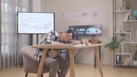 asian male sleeping while working on a car design sketch on table in the studio with tv and computers display 3d electric car model