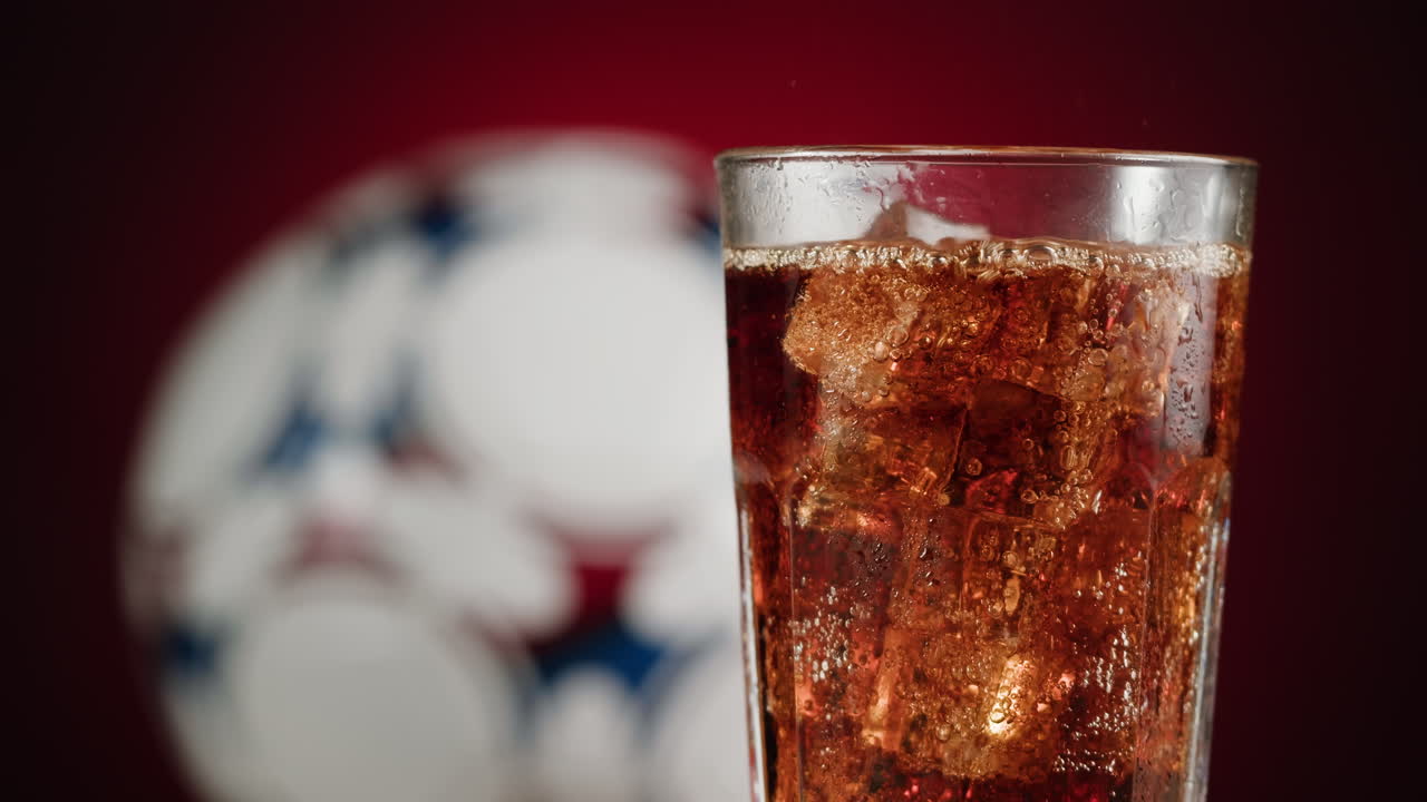 Soda in a glass with ice cubes and a soccer ball in the background