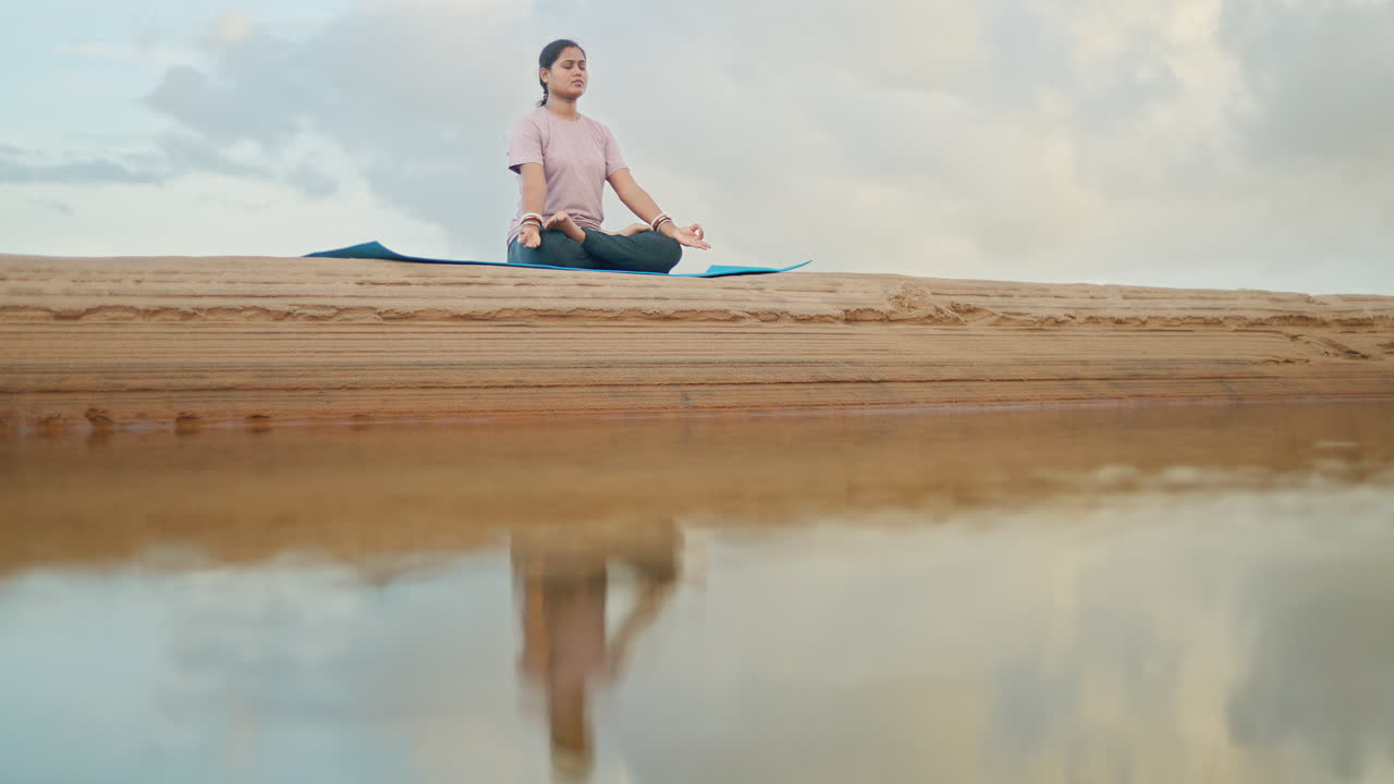 Peaceful woman sitting in lotus pose beside reflective water in a natural riverside environment in the morning