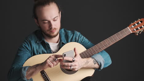 caucasian young man scrolling on smartphone while holding guitar.