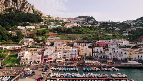 An-ascending-aerial-shot-from-the-boats-in-busy-Marina-Grande,-over-stunning-cliffs-and-winding-roads-up-to-the-boutique-shops,-fashionable-cafés-and-luxury-hotels-of-Capri-Town-in-Campania,-Italy