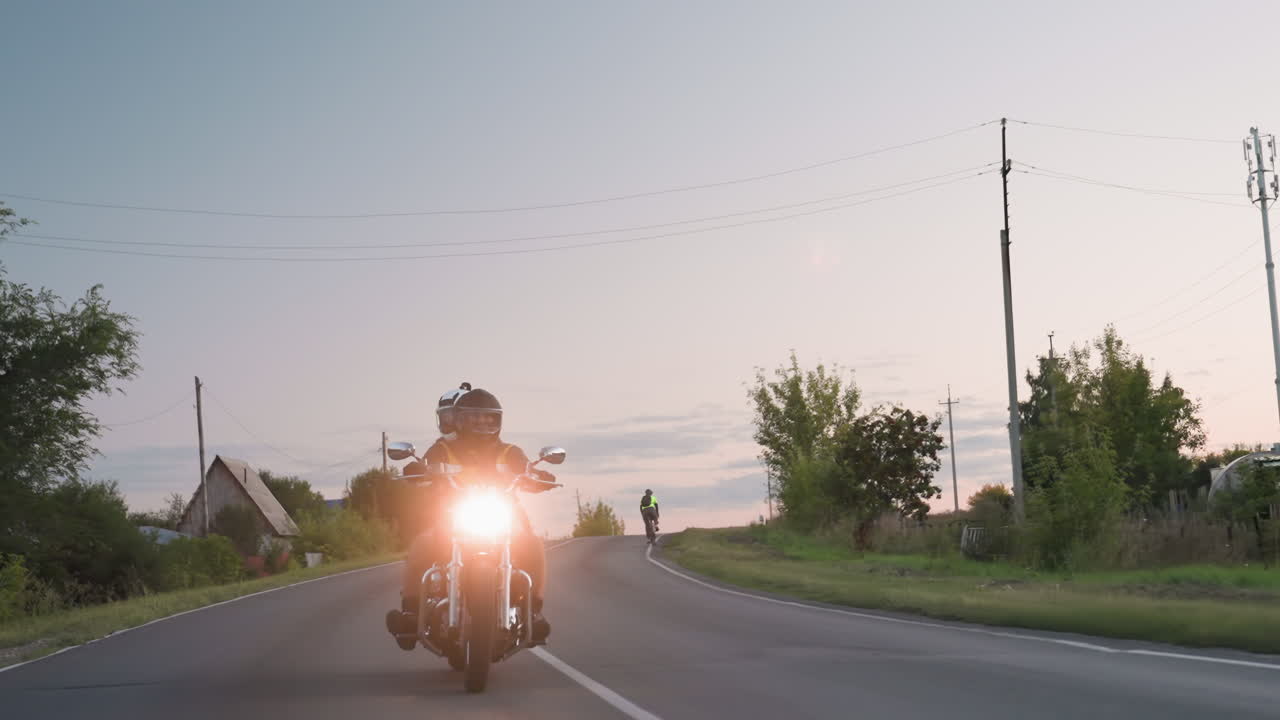 Two friends wearing helmets ride motorbike on evening countryside road, headlight glowing brightly as they make bend past trees and rural background, soft sunset sky