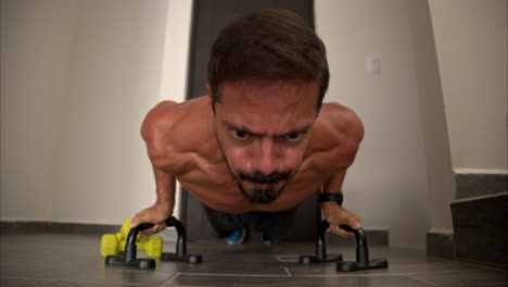 Cierre-En-Cámara-Lenta-De-Un-Hombre-Latino-Hispano-En-Forma-Entrenando-En-Casa-Haciendo-Flexiones-Usando-Barras-De-Soporte