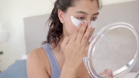 Focused-biracial-teenage-girl-putting-on-under-eye-masks-looking-in-handheld-mirror,-slow-motion