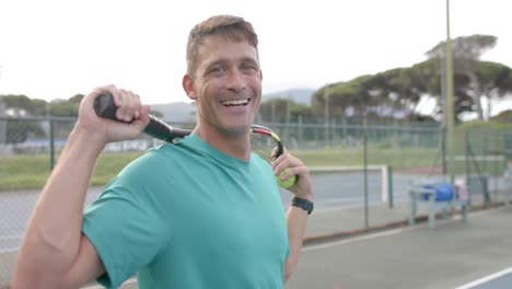 retrato de un feliz jugador de tenis caucásico que sostiene una raqueta en una cancha al aire libre, cámara lenta