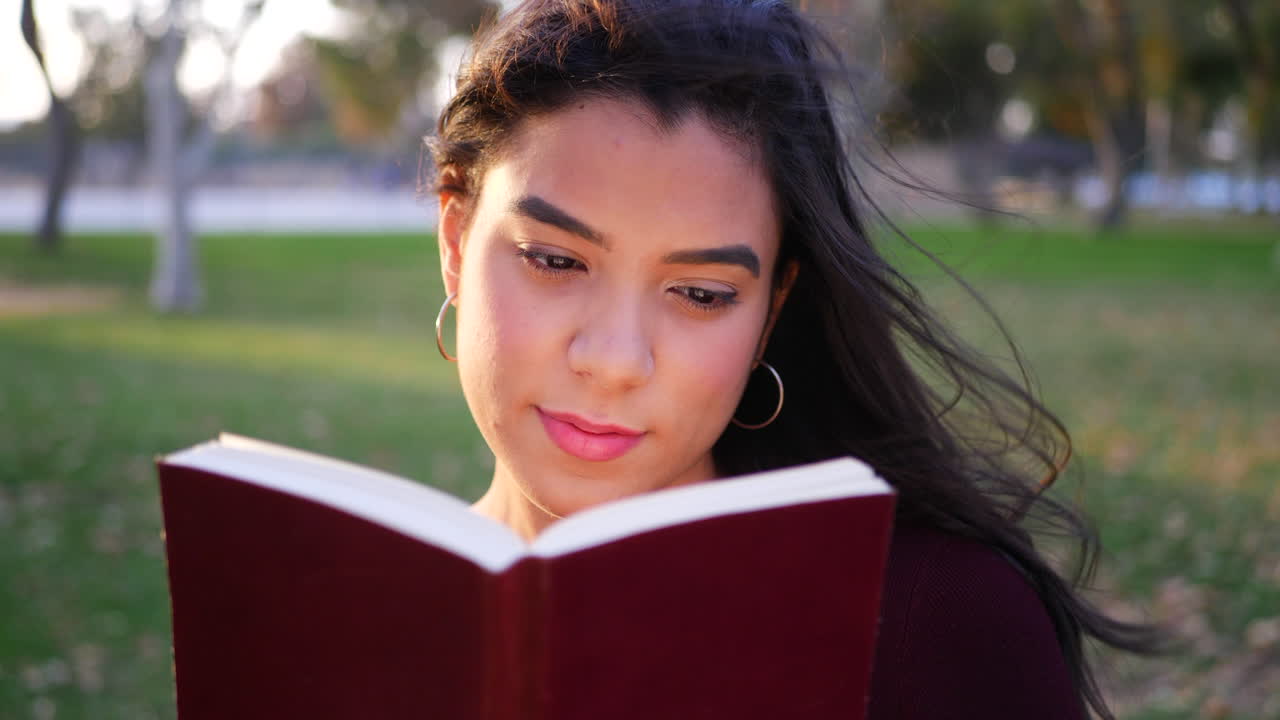 Close up on a young hispanic woman college student reading the pages of a red story book or novel outdoors SLOW MOTION