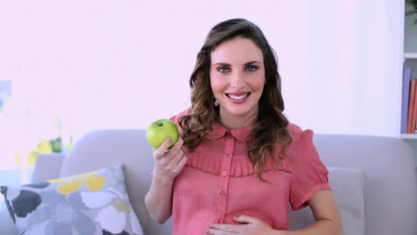 bonita modelo embarazada sentada en su sofá sosteniendo una manzana verde