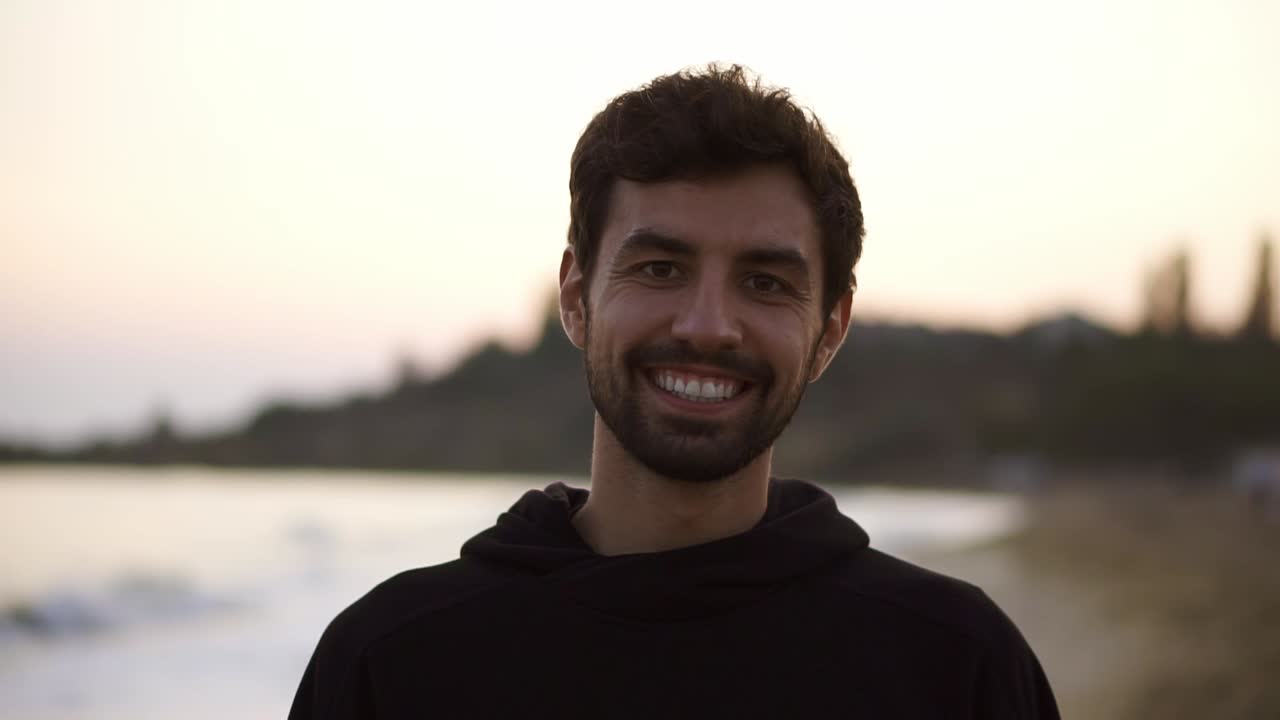 Portrait of a charming man smiling confident on calm seaside beach wearing black hoodie