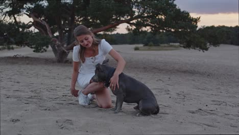 amplia vista de una joven acariciando a su perro americano staffordshire terrier en dunas de arena y mirando hacia la cámara