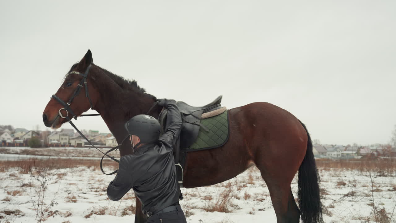 Cavaliere caucasico principiante cade mentre monta a cavallo in un campo innevato, il cavaliere con il casco viene stabilizzato dall'addestratore che assicura le redini e conduce il cavallo dolcemente. La scena mostra un paesaggio invernale, un cavallo marrone, una sella.