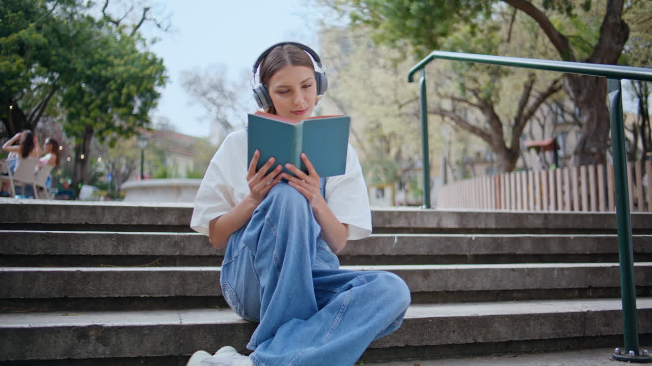 Une jeune fille avec un casque audio apprécie la lecture, assise sur des marches en béton dans un parc. Une femme qui lit