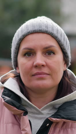 mujer confiada en chaqueta rosa y gorra gris mirando a la cámara con una expresión serena, de pie al aire libre con el follaje de otoño borroso, la casa y el jardín en el fondo