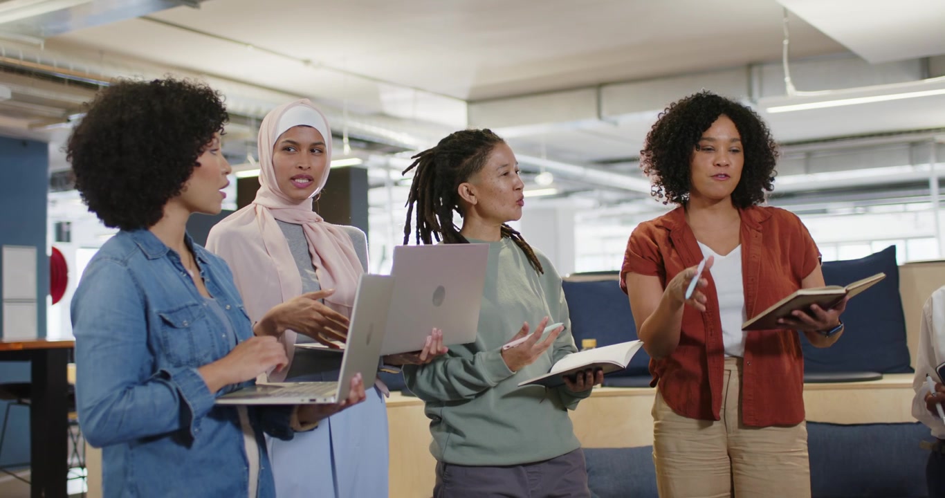 Diverse team open stand-up, woman rust top with notes, speaking, team noting laptops