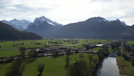 Drohnenclip,-Der-An-Einem-Hellen,-Sonnigen-Frühlingstag-Eine-Alpine-Bergkette-Mit-Schneebedeckten-Bergen-Und-Flachem-Tiefland-Darunter-Zeigt