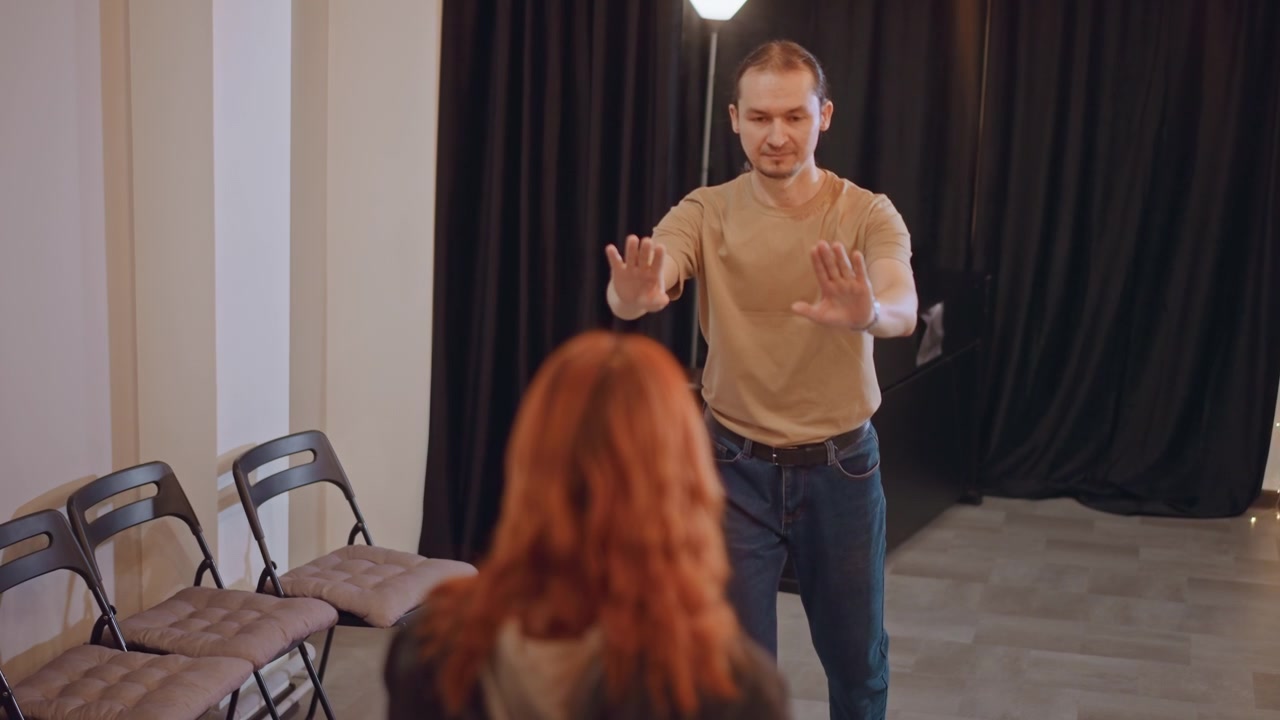 Campo lungo di un coach di recitazione maschio bianco che guida uno studente in un piccolo studio con tende nere; l'istruttore usa le mani espressive per dimostrare il posizionamento scenico e gli esercizi di riscaldamento; file di sedie pieghevoli e morbide
