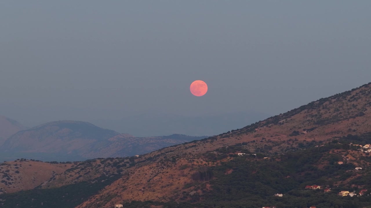 A stunning, cinematic shot of a massive pink full moon glowing in the hazy twilight sky, slowly shifting over the rugged mountain ridges of Corfu Island, Greece, during a serene summer evening.