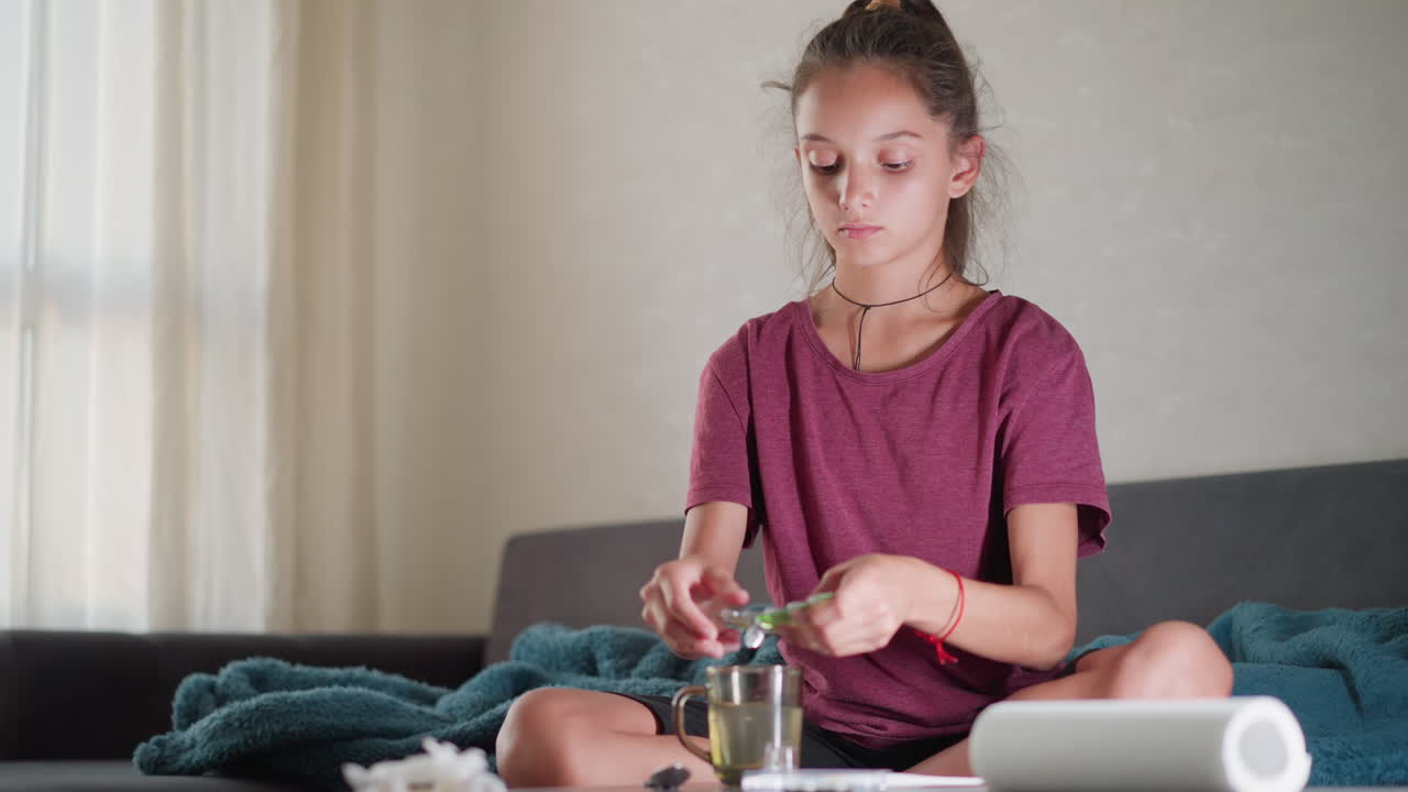 Focused girl seated on couch drops drug into glass cup of water and prepares to stir it with spoon while tissue and medical items are on table in warm home setting