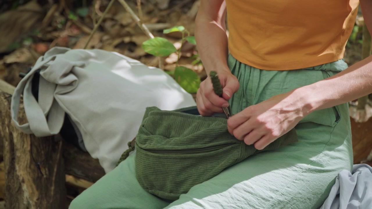 Primo piano di una donna seduta all'aperto su legno, che indossa un top arancione e pantaloni verdi. La donna interagisce con gli occhiali mentre li ripone nella sua piccola borsa scura, accanto a uno zaino di colore chiaro.