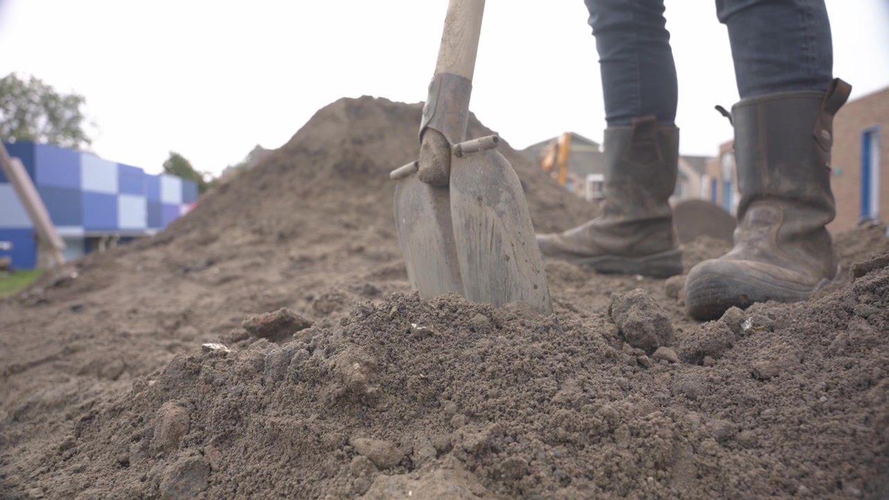 A low angle a worker standing on sand pile, shoving shovel into loose soil on a construction site, rubber boots and jeans visible.