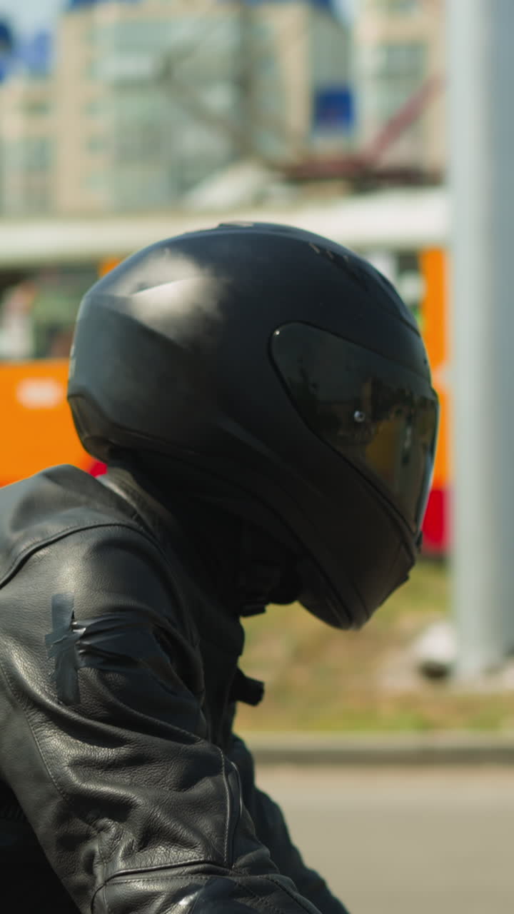 Motorcyclist man in black helmet with closed visor and leather jacket rides modern cycle along large city street on sunny day side view slow motion