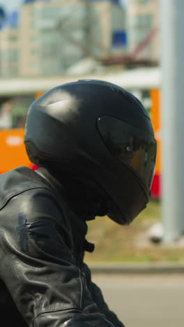 motorcyclist man in black helmet with closed visor and leather jacket rides modern cycle along large city street on sunny day side view slow motion