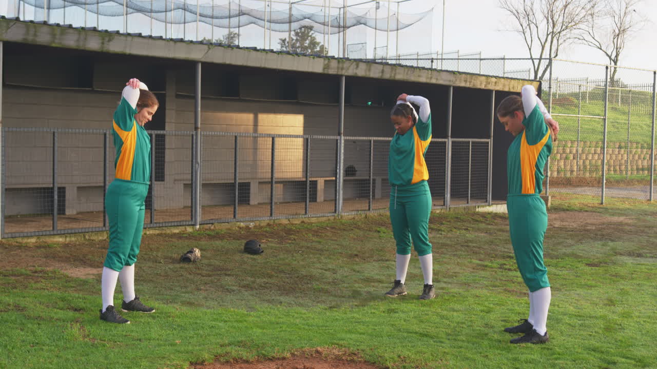 Multiracial female baseball players wearing green uniforms warming up and stretching arms on a pitch