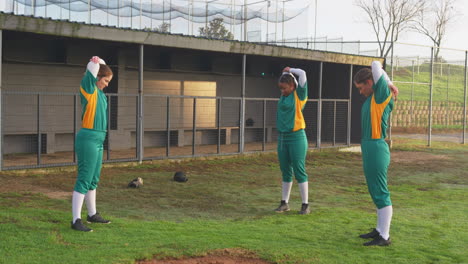 multiracial female baseball players wearing green uniforms warming up and stretching arms on a pitch