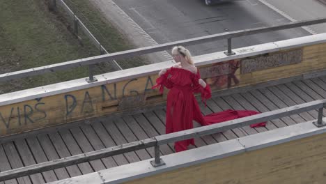 Aerial-shot-of-a-model-in-red-dress,-standing-on-wooden-bridge-with-a-road-behind-her