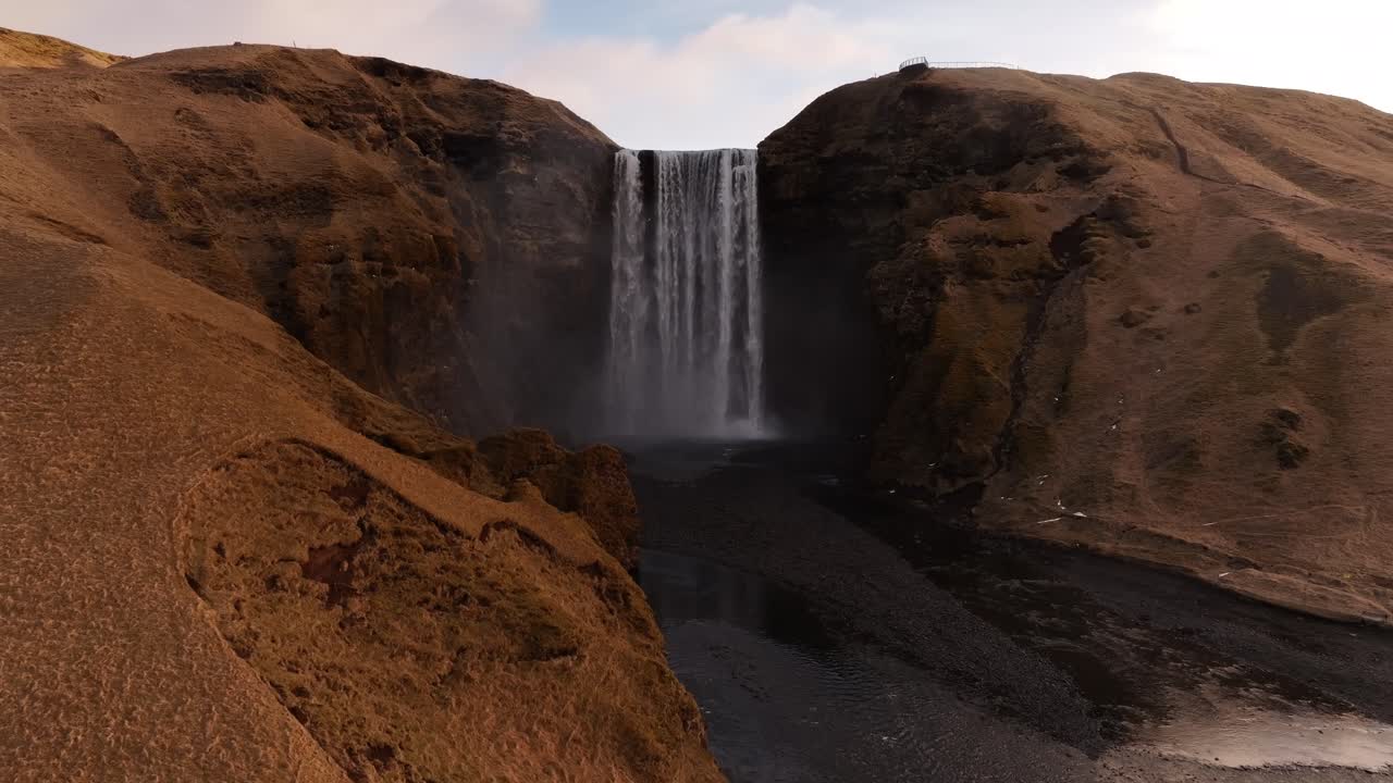 Slow cinematic drone push-in toward Skógafoss waterfall in Iceland, landscape with mossy volcanic cliffs, black riverbed and epic mist.